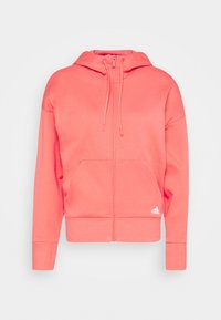 Felpa con zip color corallo realizzata in tessuto morbido, dotata di tasca a marsupio, cappuccio regolabile con cordino e logo Adidas discreto in basso a sinistra.