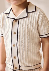 Strikket cremefarvet skjorte med strukturerede lodrette striber, sort kant, korte ærmer og seks træknapper foran.