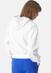 Felpa bianca con cappuccio e vestibilità rilassata, con polsini a costine, abbinata a pantaloni blu che mostrano un dettaglio della tasca posteriore. Tessuto morbido con una superficie liscia.