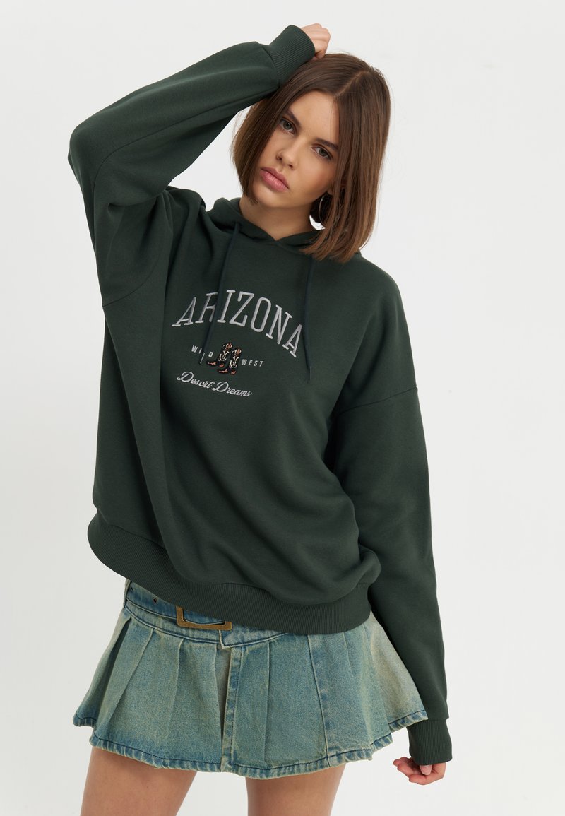Diep groene hoodie met een logo waarop "ARIZONA Wild West Desert Dreams" staat, gecombineerd met een lichtblauwe denim plooienrok.