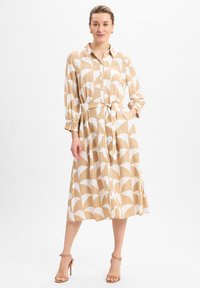 Lange, knoopsluiting jurk van lichtgewicht stof met een beige achtergrond en witte golfpatronen. Beschikt over een riem en elastische mouwmanchetten.