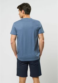 Modrá krátká košile s kulatým výstřihem, v kombinaci s námořnickými šortkami. Tkanina vypadá měkce s hladkým povrchem. Zobrazený zadní pohled.