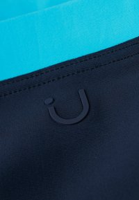 Marineblauer Stoff mit einer strukturierten Oberfläche, versehen mit zwei aquafarbenen Streifen und einem dunklen Logo-Patch an einer genähten Naht.