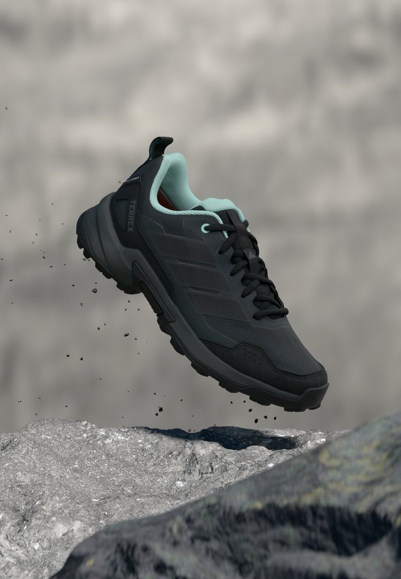 Μαύρο αθλητικό παπούτσι με ανοιχτό μπλε λεπτομέρειες, με υφή στο επάνω μέρος, κορδόνια και ανθεκτική σόλα, αιωρούμενο πάνω από μια πετρώδη επιφάνεια.