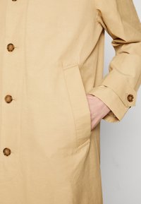Beige trenchcoat met schildpadmotief knopen, zijzak en manchetband, hand in de zak gestoken.