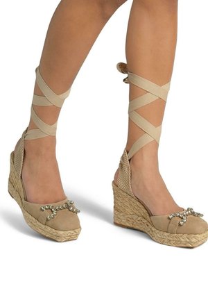 Le espadrillas beige con tacco wedge presentano una suola in juta intrecciata, una parte superiore in tessuto testurizzato e un dettaglio di fiocco decorativo. Le cinghie si avvolgono attorno alle caviglie.