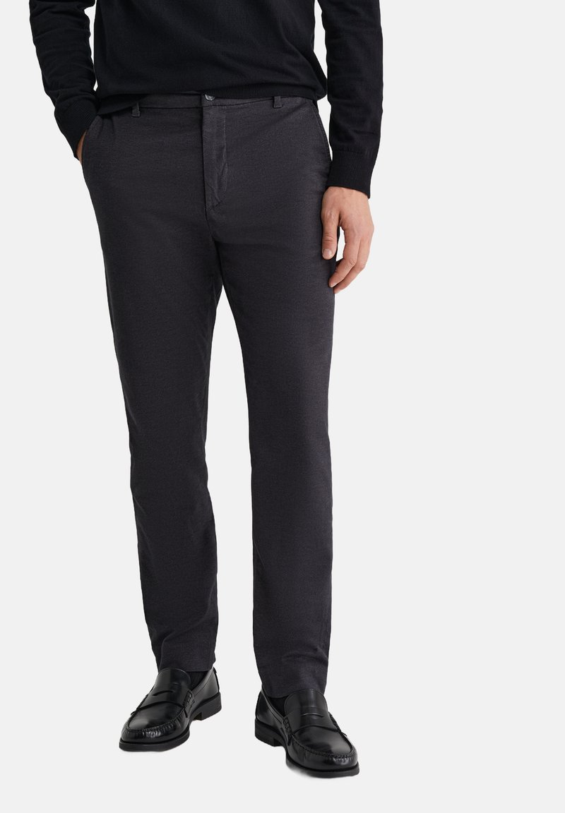 Muž v tmavě šedých úzkých kalhotách, černých mokasínách a černém dlouhém rukávu s rukama v kapsách na bílém pozadí.