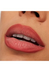 Matowe usta w odcieniu terakoty o gładkiej teksturze, subtelnie zdefiniowanym kształcie i widocznych drobnych liniach. Tło przedstawia neutralny odcień skóry.