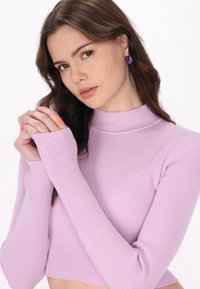 Maglione a collo alto a coste di un viola chiaro con maniche lunghe; design aderente che mette in risalto la texture. Le mani del modello sono adornate con anelli colorati.