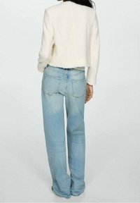 Kvinde iført lyseblå lige ben jeans og en hvid, struktureret langærmet cropped jakke, stående med ryggen til mod en ensfarvet baggrund.