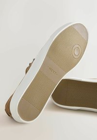 Semelles de chaussures marron avec bords blancs, une semelle tournée vers le haut montrant le texte "NEXT" et un motif de grip texturé sur caoutchouc beige.