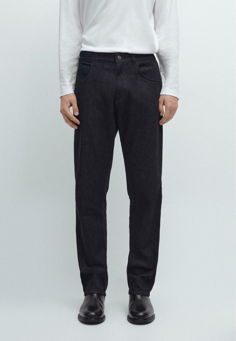 Jeans in denim scuro con vestibilità dritta, dotati di chiusura con bottone, tasche classiche e dettagli di cucitura sottili. Indossati con una camicia bianca a maniche lunghe.