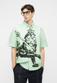 Jeune homme portant des lunettes et une chemise à manches courtes vert clair avec un grand motif graphique noir et un pantalon noir.