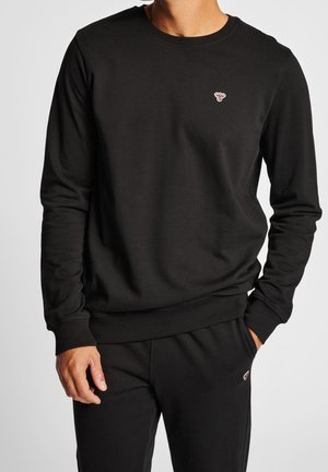 Mann trägt schwarzen Langarm-Sweatshirt und passende schwarze Hose mit kleinen bestickten Bienen-Logos auf Brust und Oberschenkel.