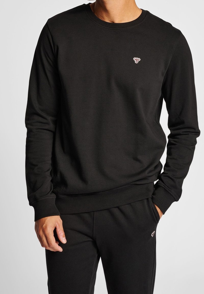 Homme portant un sweat-shirt noir à manches longues et un pantalon noir assorti avec de petits logos d’abeille brodés sur la poitrine et la cuisse.