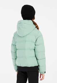 Grüne gepolsterte Jacke mit Kapuze, gerippten Bündchen und Saum, getragen mit einer schwarzen Strickmütze. Rückansicht, die die glatte Textur und die kastige Form zeigt.