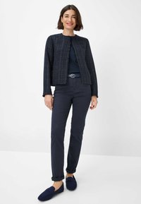 Texturierte marineblaue Jacke mit einem kurzen Design, kombiniert mit marineblauen Slim-Fit-Hosen und dunklen Slippern. Eng anliegende Silhouette, minimaler Hardware.