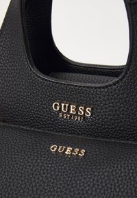 Crna torba s teksturama i glatkom završnom obradom. Sadrži zlatni logo "GUESS" i godinu osnivanja. Geometrijskog oblika s dva ručka.