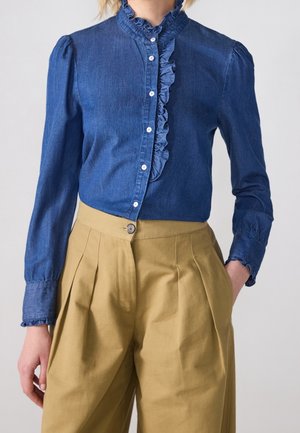 Mujer que lleva una blusa azul de mezclilla con botones y volantes, combinada con pantalones beige plisados de talle alto, una mano en el bolsillo.