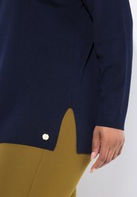 Marineblaue Sweater mit seitlichem Schlitz und goldenem Emblem, getragen über senfgelben Hosen, zeigt eine entspannte Hand an der Seite.
