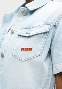 Lyseblå denimjakke med metal knapper og en brystlomme foran broderet med røde "RBR" bogstaver.