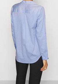 Camicia azzurra chiara con un motivo discreto, maniche lunghe e orlo curvo. La vista posteriore mostra un yoke e una pince centrale.