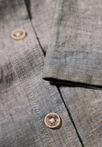 Close-up van een grijze linnen overhemd met een gestructureerde stof, voorzien van een ronde, lichtgekleurde knop en zichtbare stikdetails.