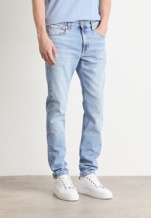 Mies, jolla on päällään vaaleansiniset slim fit -farkut ja valkoiset tennarit, seisoo kädet taskuissa yksiväristä vaaleaa taustaa vasten.