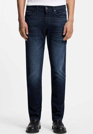 Homme portant un jean slim bleu foncé et des chaussures en cuir noires, debout les mains détendues le long du corps sur fond blanc.