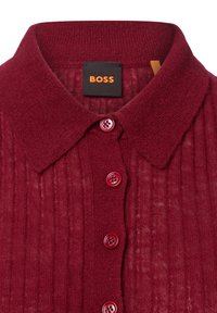 Chemisier en tricot rouge côtelé avec un col classique et cinq boutons assortis. L'étiquette indique "BOSS" sur fond noir. Détail de tissu texturé visible.