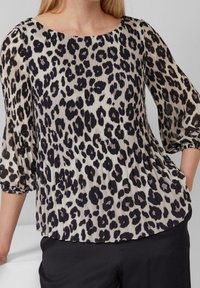 Blouse beige clair à imprimé léopard noir. Texture plissée, encolure ronde et manches ballon trois-quarts. Remarque : pantalon noir visible.