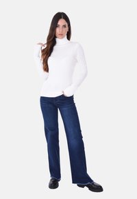 Maglione bianco a collo alto, maniche lunghe aderenti, abbinato a jeans a zampa d'elefante blu scuro, con orlo sfilacciato e scarpe nere chunky.