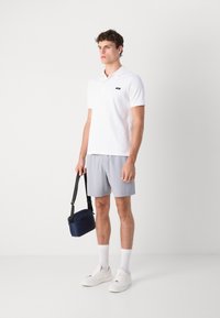 Hvitt bomullspolo, lysegrå shorts, hvite sneakers og en mørkeblå skulderveske. Modellen står mot en ensfarget bakgrunn.