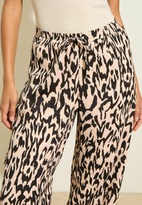 Kvinna klädd i beige och svart leopardmönstrade byxor med dragsko i midjan, kombinerat med en ljusbeige ärmlös topp.
