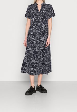 Kobieta w czarnej sukience midi w białe grochy z krótkimi rękawami, zestawiona z masywnymi czarnymi mokasynami, stojąca na jednolitym tle.