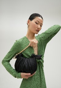 Bolso negro texturizado con diseño fruncido, cadena dorada y acentos de lazo, sostenido por una persona que lleva un vestido verde con estampado.