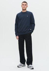 Navy blue crew neck sweatshirt in cotton blend, featuring a small emblem on the chest, paired with black straight-leg trousers and gray sneakers.