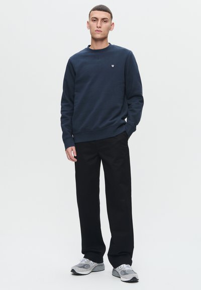 Sweat-shirt bleu marine à col rond en mélange de coton, avec un petit emblème sur la poitrine, associé à un pantalon noir à jambe droite et des baskets grises.