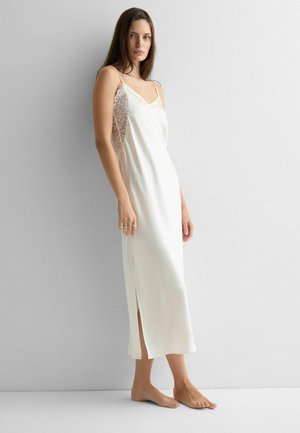 Frau, barfuß stehend, trägt ein langes weißes Nachthemd mit seitlichen Spitzenpartien und dünnen Schulterträgern vor einem schlichten grauen Hintergrund.