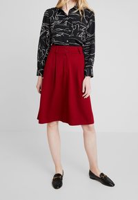 Svart blus med vitt linjemönster, röd A-linjeformad kjol med veck och knappar, samt svarta loafers med guldaccenter.