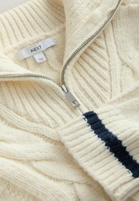 Cremfarbener Kabelstrickpullover mit silbernem Reißverschluss und navyfarbener Streifen am Bündchen. Das Etikett zeigt "NEXT" und die Pflegehinweise sind sichtbar.