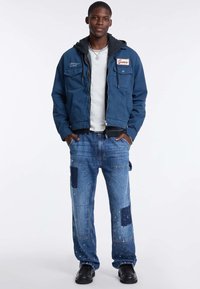 Giacca navy con cappuccio, tasche frontali e logo "Guess" ricamato; abbinata a jeans patchwork blu chiaro e scarpe nere.