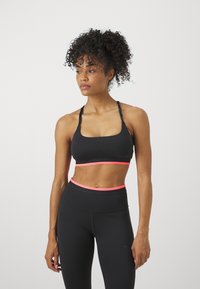 Mujer atlética con cabello rizado que lleva un sujetador deportivo negro y leggings con ribetes rosas, mirando hacia un lado contra un fondo blanco liso.
