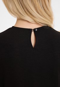Haut noir avec une découpe en forme de clé à l'arrière, maintenue avec un bouton noir. Le matériau semble doux et flexible.