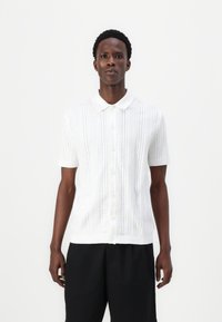 Balts īsajiem piedurknēm polo džemperis, kas izceļas ar adītu rakstu ar vertikālām līnijām un caurumiem; pogas priekšpusē; maiga tekstūra.