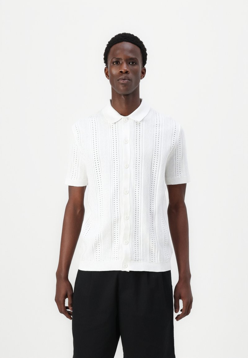 Balts īsajiem piedurknēm polo džemperis, kas izceļas ar adītu rakstu ar vertikālām līnijām un caurumiem; pogas priekšpusē; maiga tekstūra.