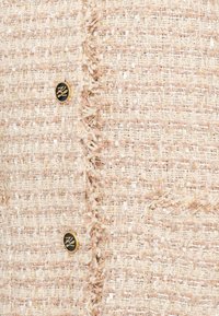 Bežová texturovaná tkanina z tweedu s třepeným okrajem a zlatým knoflíkem se zabudovaným logem.