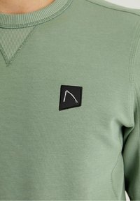 Maglione a maniche lunghe di un verde chiaro a coste, con una piccola toppa nera che presenta un motivo astratto bianco sul petto.