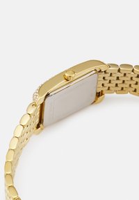 Goldfarbene Uhr mit rechteckigem Gehäuse, Edelstahl-Mesh-Armband, strukturiertem Rand und kleiner Krone, mit transparentem Rückenteil.