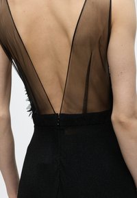 Πλάτη ατόμου που φοράει μαύρο φόρεμα με βαθύ V-σχήματος διαφανές δίχτυ στην πλάτη και εφαρμοστό ζωστικό, που δείχνει γυμνό δέρμα μέσα από το ύφασμα.